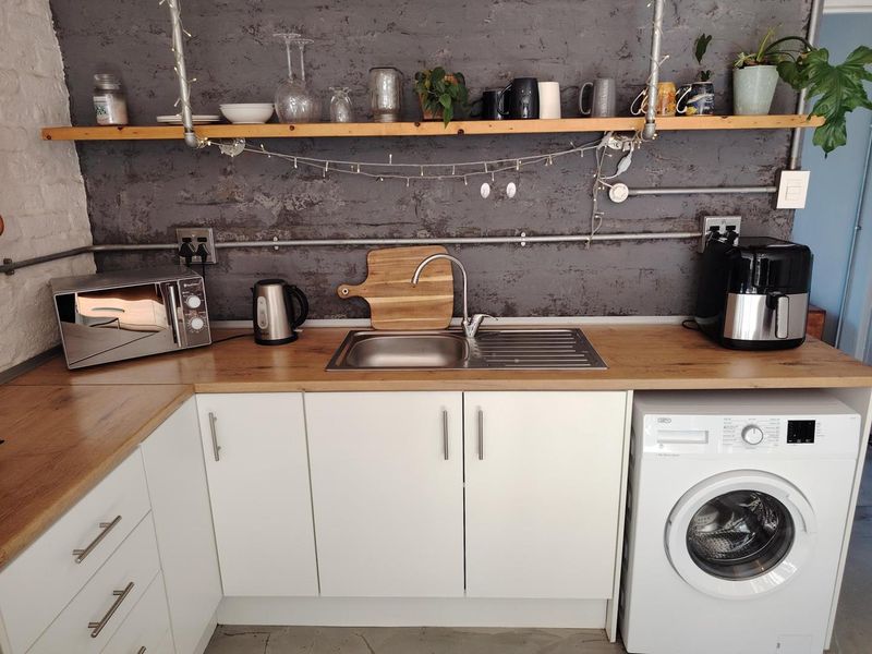 A compact kitchen area featuring white cabinetry, a wooden countertop, stainless steel sink, and industrial-style exposed shelving against a textured grey wall.