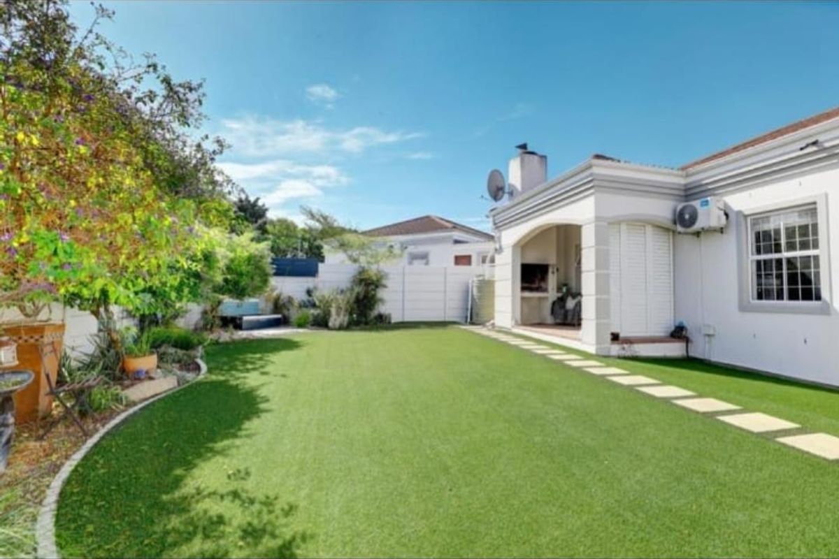 A well-maintained rear garden featuring an artificial lawn, paved walkway, and a covered patio area with a built-in braai.