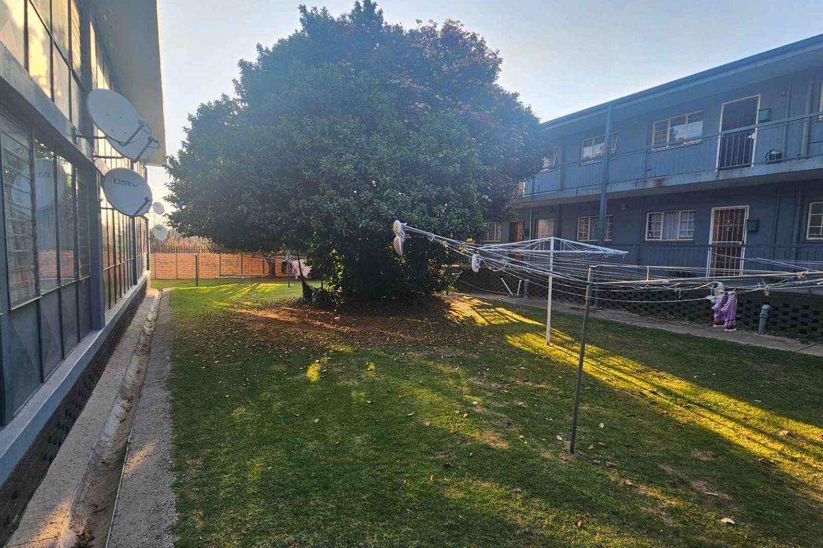 Rear exterior view of the apartment complex featuring a communal lawn area, mature tree, and building facade with security features.