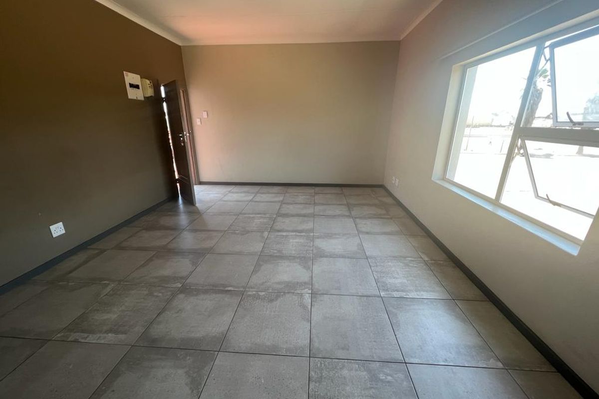 An empty, open-plan living area featuring large grey floor tiles, neutral wall tones, and a large window providing natural light.