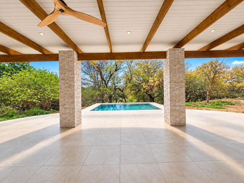 A spacious covered patio featuring stone-clad pillars, timber ceiling beams, and recessed lighting, overlooking a private swimming pool and natural landscape.