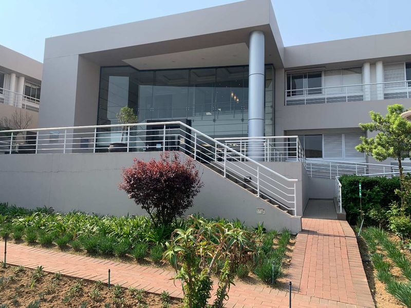 Modern office building exterior featuring a glass facade, elevated entrance with stairs and ramp access, and landscaped garden area.