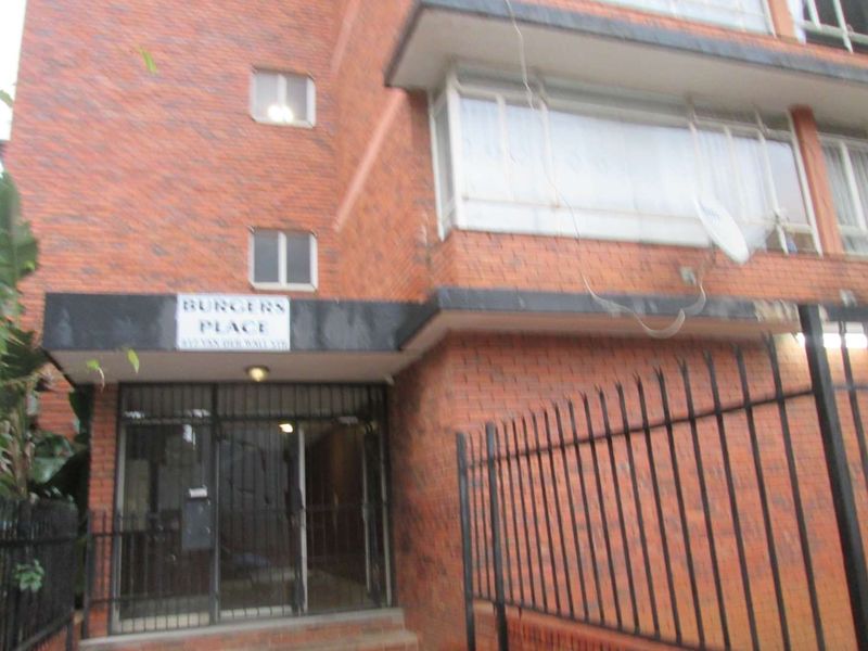 Front entrance view of the brick apartment building known as Burgers Place, featuring a secure gated entry and metal perimeter fencing.