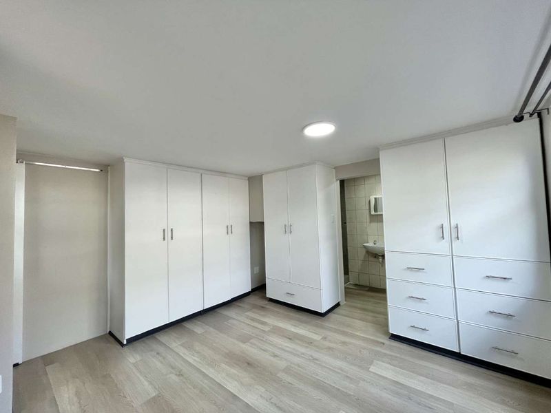A bright, empty bedroom featuring extensive white built-in cabinetry, modern light-toned laminate flooring, and direct access to an en-suite bathroom.