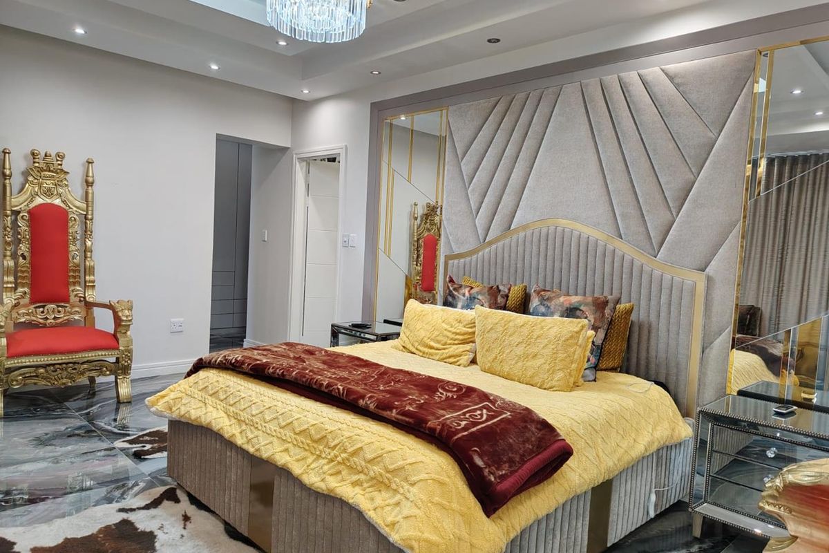 A luxurious bedroom featuring a large upholstered headboard, recessed ceiling with downlights, a crystal chandelier, and polished marble flooring.