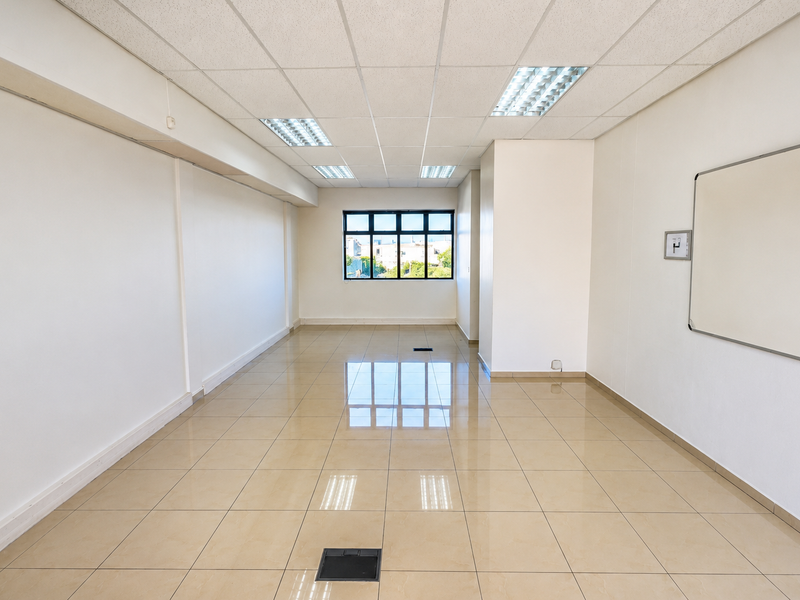 A bright, empty office space featuring polished tiled flooring, a suspended ceiling with recessed lighting, and a wall-mounted whiteboard.
