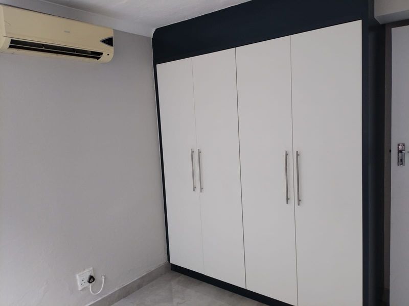 A clean bedroom interior featuring a modern white built-in wardrobe, a wall-mounted air conditioning unit, and neutral grey wall finishes.
