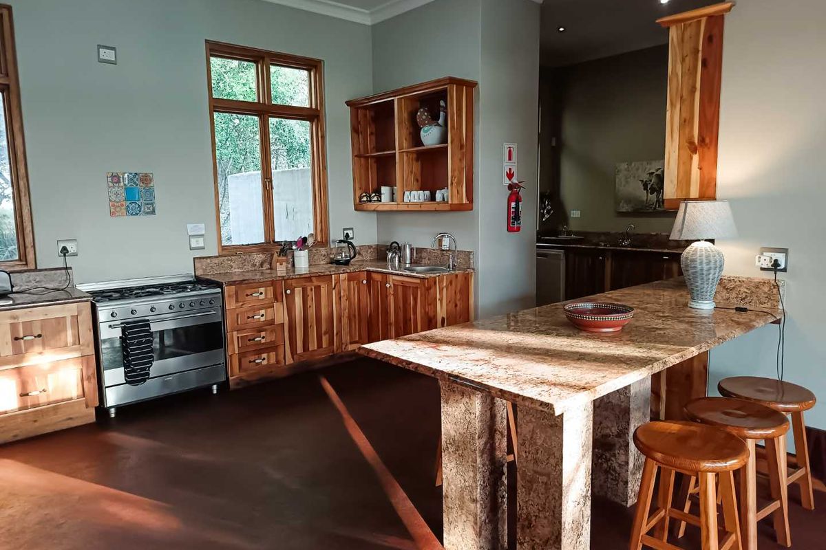 A rustic-style kitchen featuring wooden cabinetry, granite countertops, a central island, and a stainless steel gas stove, with an adjacent scullery area.