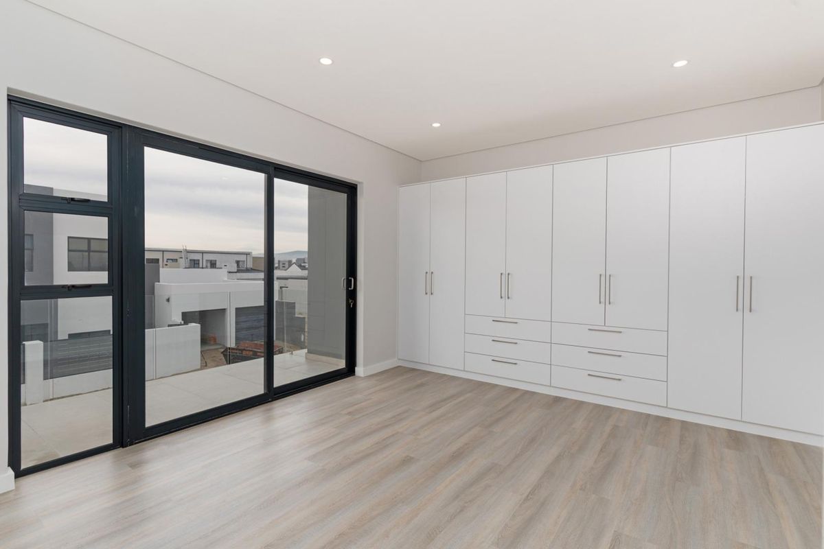 A bright, modern bedroom featuring extensive floor-to-ceiling built-in cabinetry, light wood-look flooring, and large sliding glass doors opening to a private balcony.
