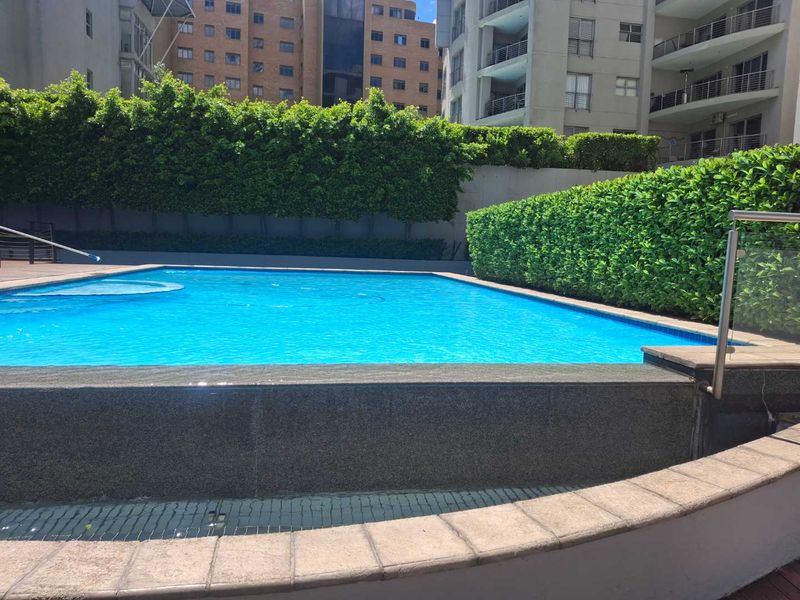 A well-maintained communal swimming pool area featuring a tiled coping, stainless steel safety railing, and established privacy hedging within an apartment complex.