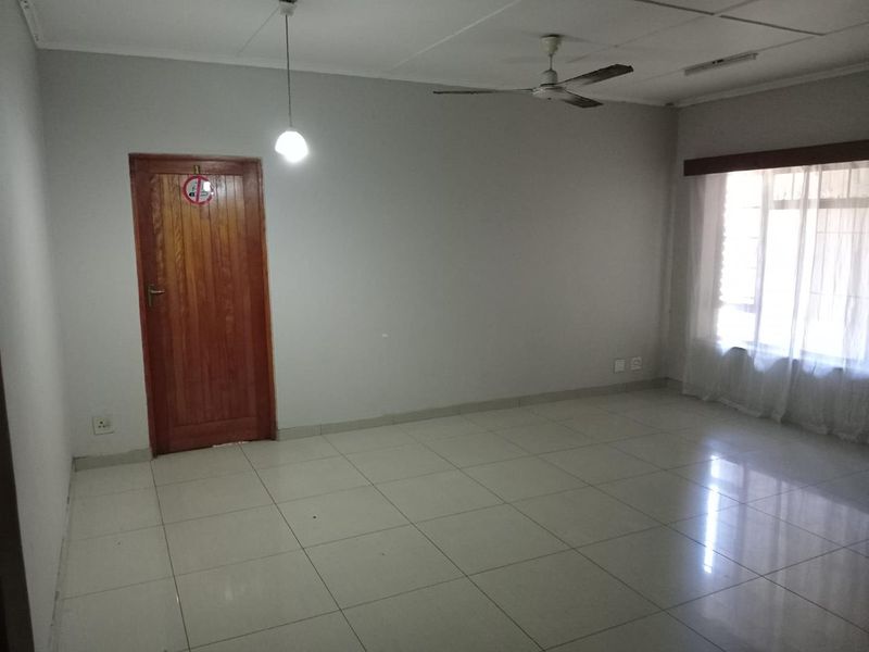 A spacious, empty living area featuring neutral tiled flooring, a ceiling fan, and a wooden door, offering a blank canvas for interior design.