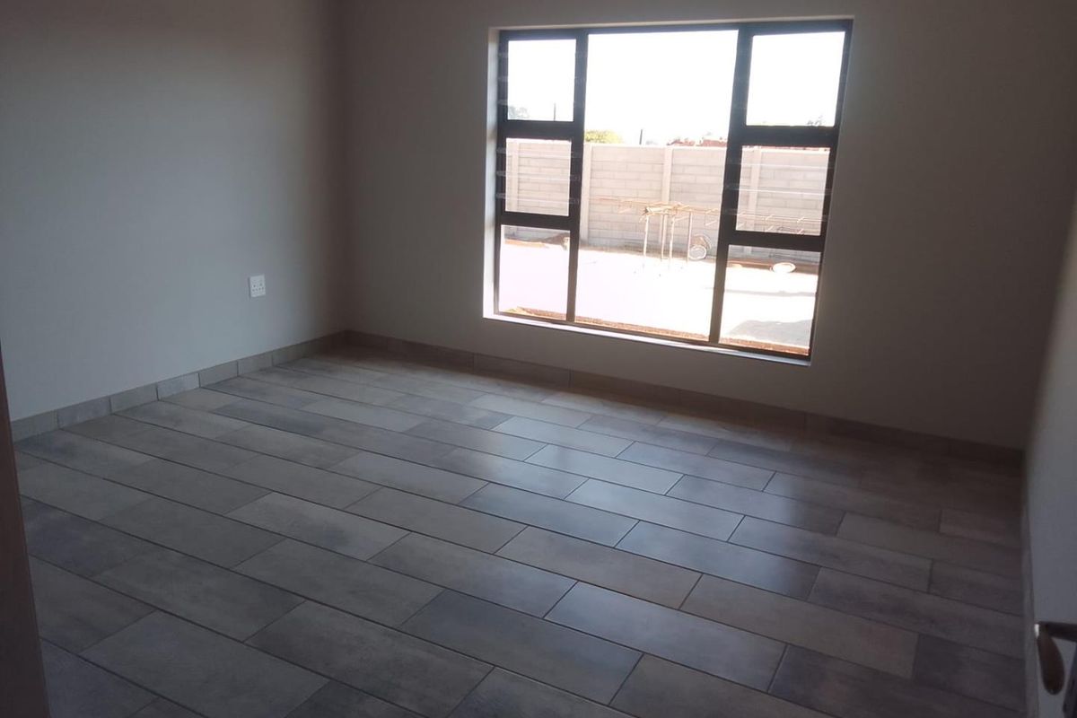 A bright, empty bedroom featuring modern grey wood-look floor tiles and a large window providing natural light.