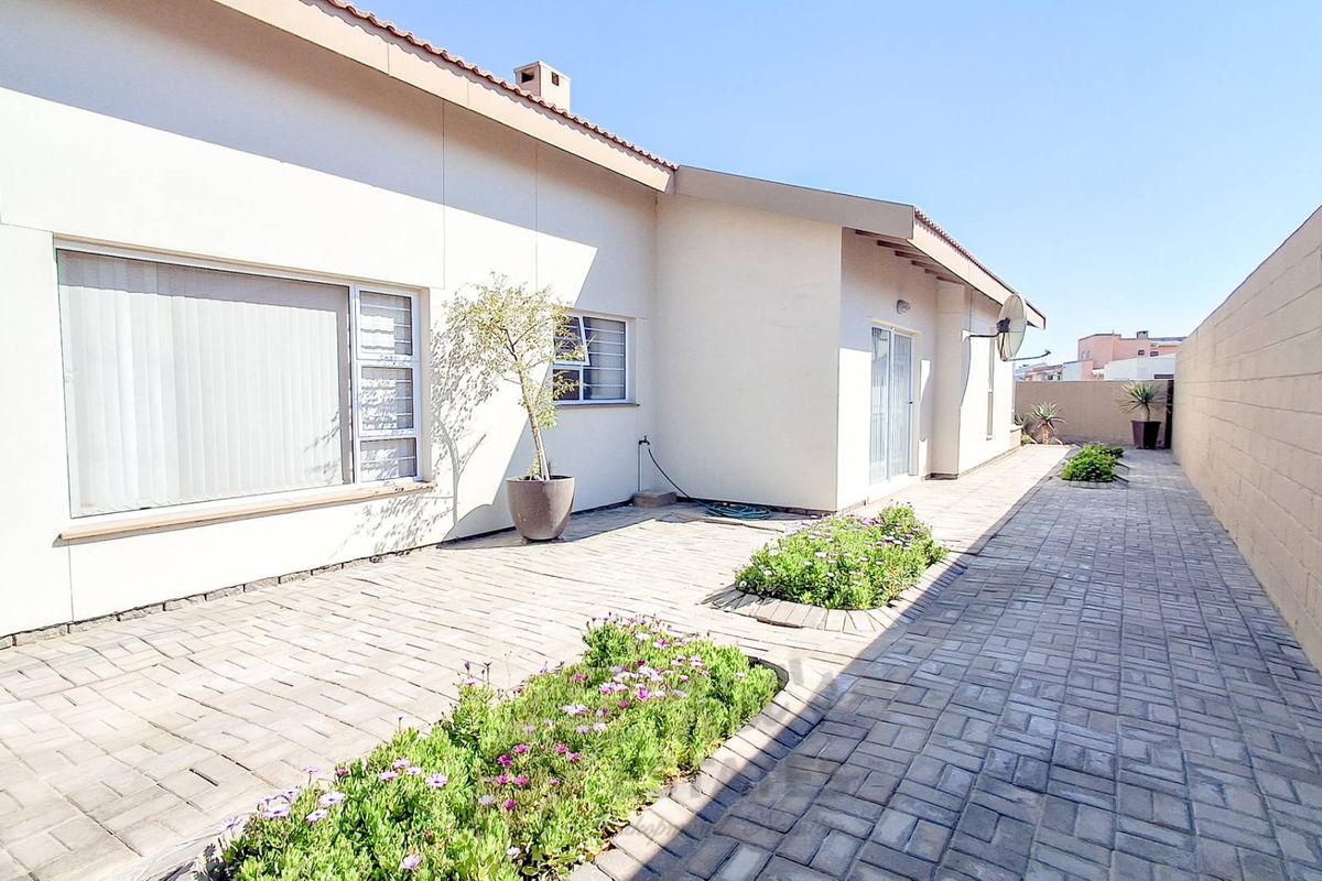 A well-maintained rear exterior view featuring paved patio areas, landscaped garden beds, and a high boundary wall for privacy.