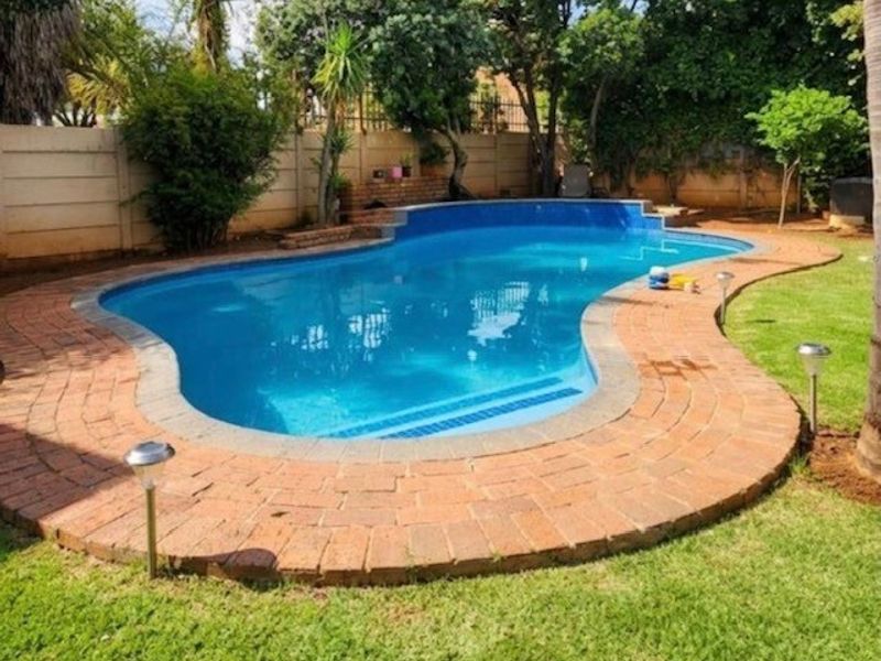 A well-maintained swimming pool area featuring a curved design, brick paved surround, and established garden landscaping with a perimeter boundary wall.