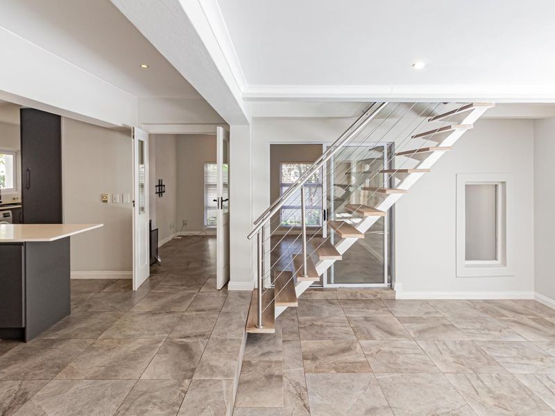 Modern open-plan living area featuring a contemporary floating staircase with glass and stainless steel balustrades, tiled flooring, and a sleek kitchen.