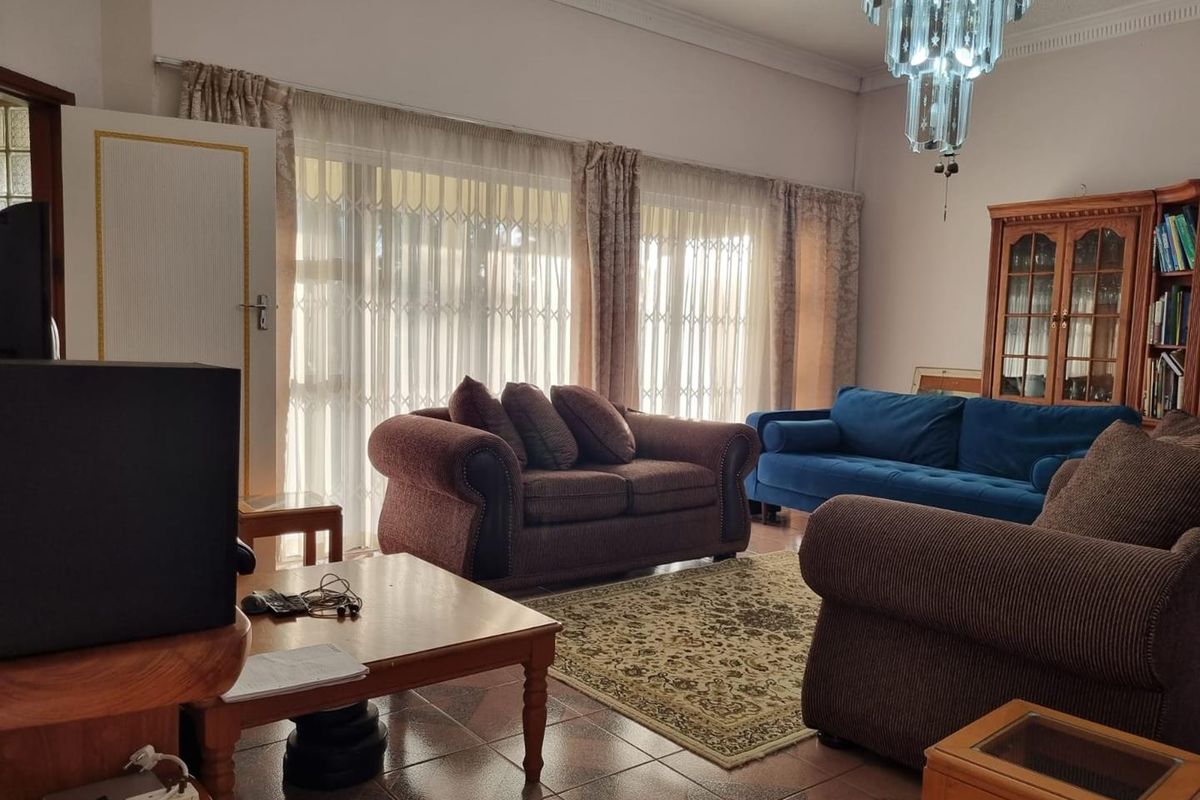 A spacious living room featuring tiled flooring, large windows with security gates, and a decorative chandelier.