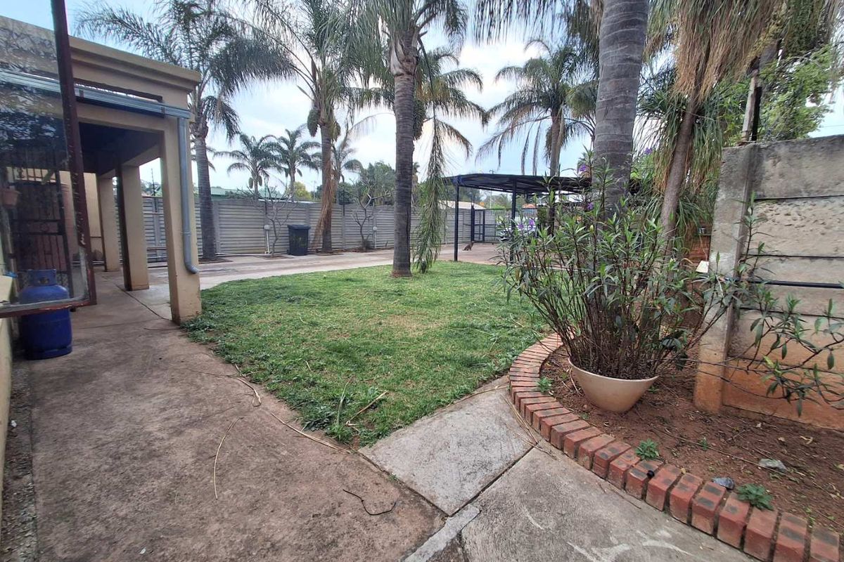 A rear garden area featuring a concrete patio, lawn, perimeter wall, and a covered carport structure.