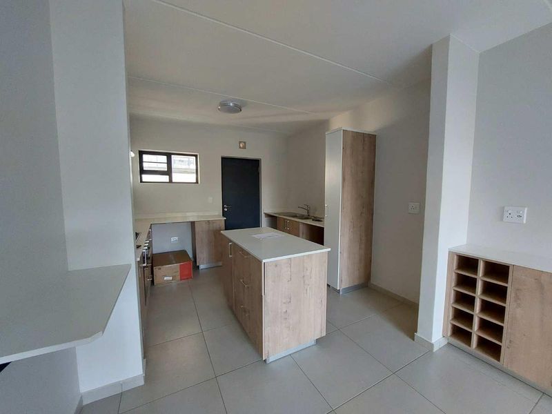 A modern, empty kitchen featuring light wood-tone cabinetry, a central island, tiled flooring, and a built-in storage unit, offering a clean and functional layout.