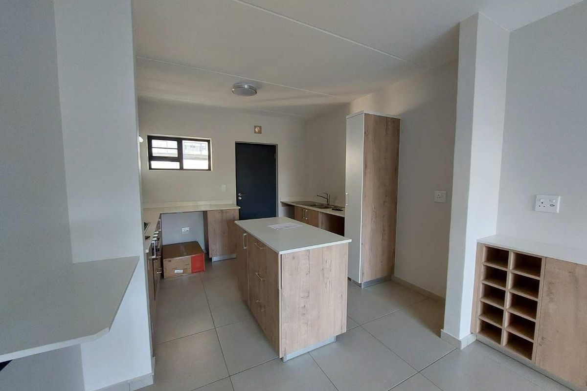 A modern, empty kitchen featuring light wood-tone cabinetry, a central island, tiled flooring, and a built-in storage unit, offering a clean and functional layout.