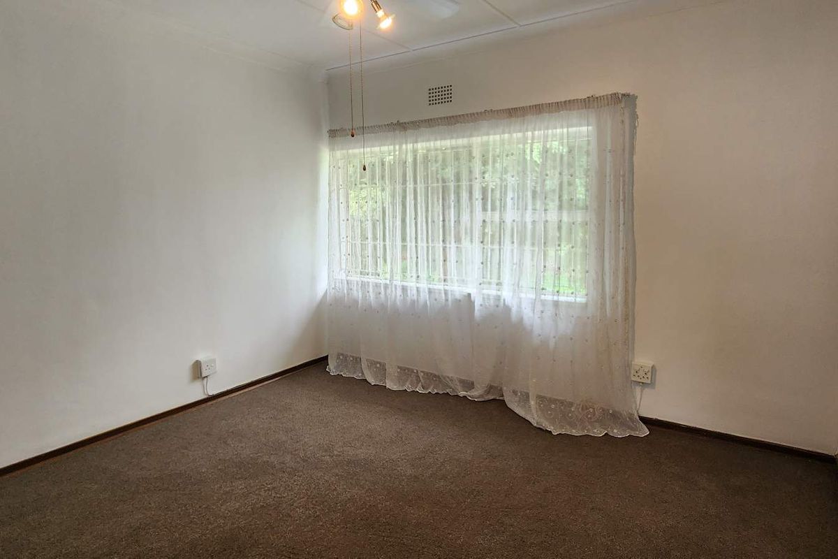 A neutral, carpeted bedroom featuring a large window with sheer curtains and a ceiling-mounted light fixture.
