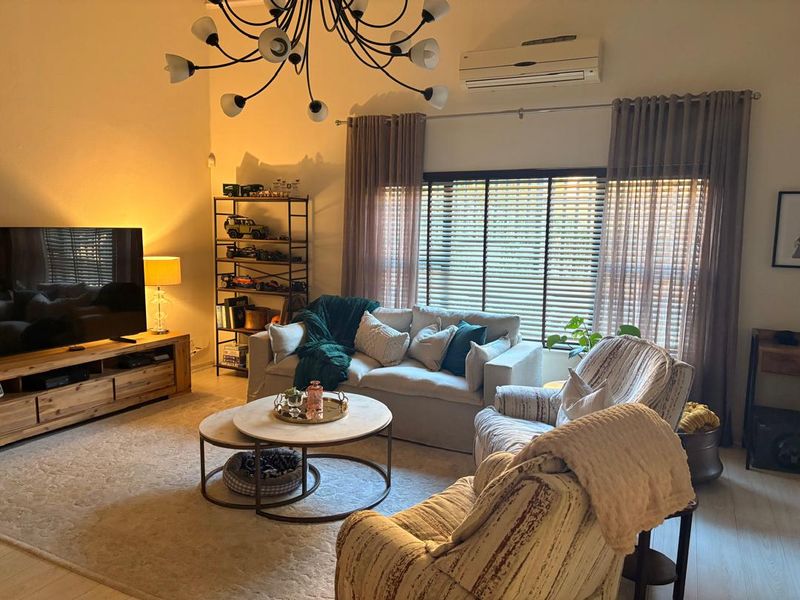 A cozy living room featuring vaulted ceilings, laminate flooring, and a wall-mounted split-system air conditioner, complemented by large windows with venetian blinds.