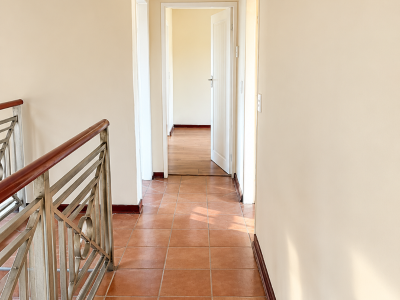 A bright, empty hallway featuring ceramic floor tiles, a metal balustrade with a wooden handrail, and neutral wall finishes leading to adjacent rooms.