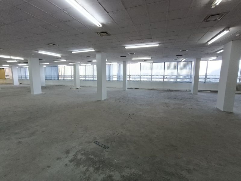 A large, open-plan office space featuring structural columns, suspended ceilings with fluorescent lighting, and expansive windows with vertical blinds.