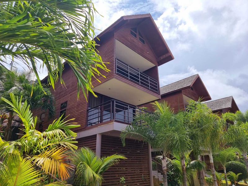 Exterior view of multi-level commercial timber-clad buildings featuring private balconies with metal railings and pitched roof architecture set within a landscaped tropical environment.