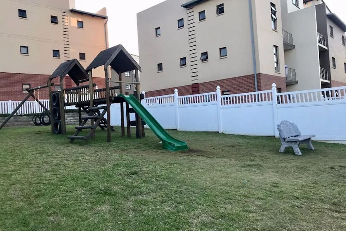 Communal garden area featuring a children's playground set and white vinyl picket fencing, situated between apartment complex buildings.