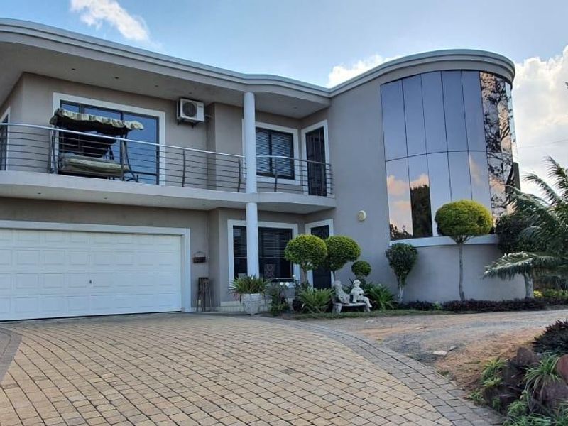 Modern two-story residence featuring a distinctive curved glass facade, a spacious balcony with stainless steel balustrades, and a paved driveway leading to a double garage.