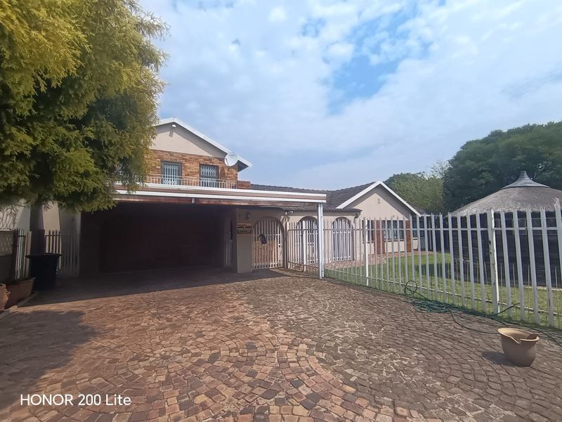 Front view of the property featuring a paved driveway, covered carport, security fencing, and a detached thatched lapa in the garden area.