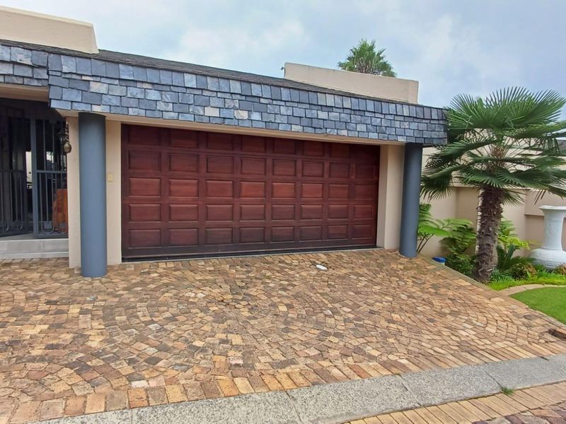 Front elevation of the property featuring a double garage with wooden door, slate roof detailing, and a paved circular pattern driveway.