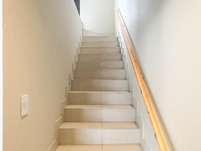 A clean, minimalist staircase featuring light-toned floor tiles and a simple wooden handrail, leading to an upper level.