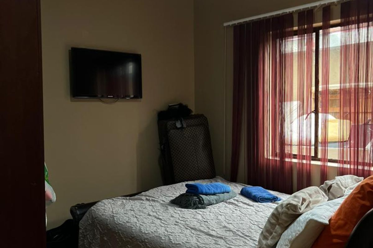 A functional bedroom featuring tiled flooring, a wall-mounted television, and a window with fitted curtain tracks.