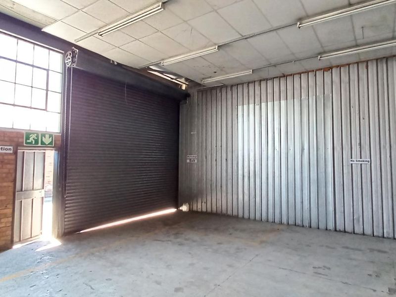 Spacious industrial warehouse unit featuring a large roller shutter door, exposed brick walls, steel cladding, and high-clearance ceilings with fluorescent lighting.