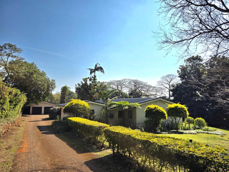 A well-maintained single-story house featuring a brick driveway, detached garage, and established garden with mature trees and manicured hedging.