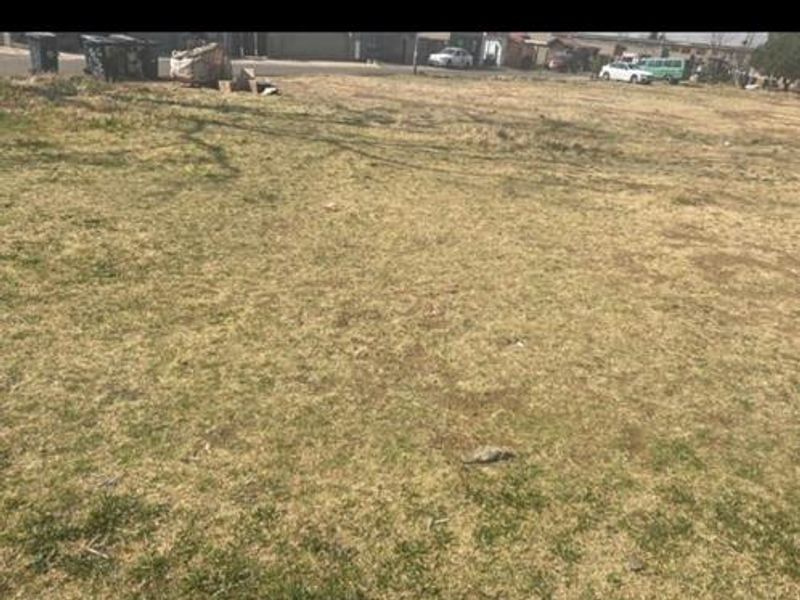 A view of a vacant residential plot of land with grass cover, showing the street frontage and surrounding neighborhood context.