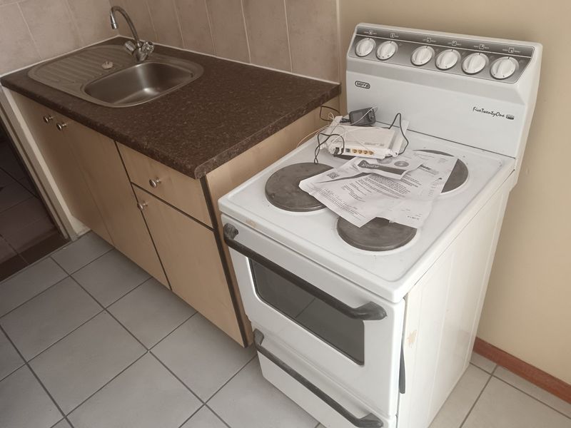 A compact kitchen area featuring a stainless steel sink, laminate countertop, tiled backsplash, and a freestanding electric stove with tiled flooring.