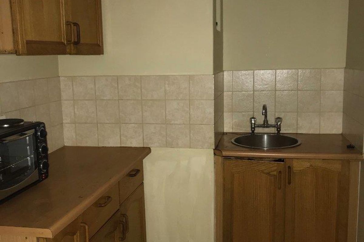 A compact kitchen featuring dated wooden cabinetry, laminate countertops, a stainless steel sink, and tiled backsplash, requiring cosmetic updates to modernize the space.