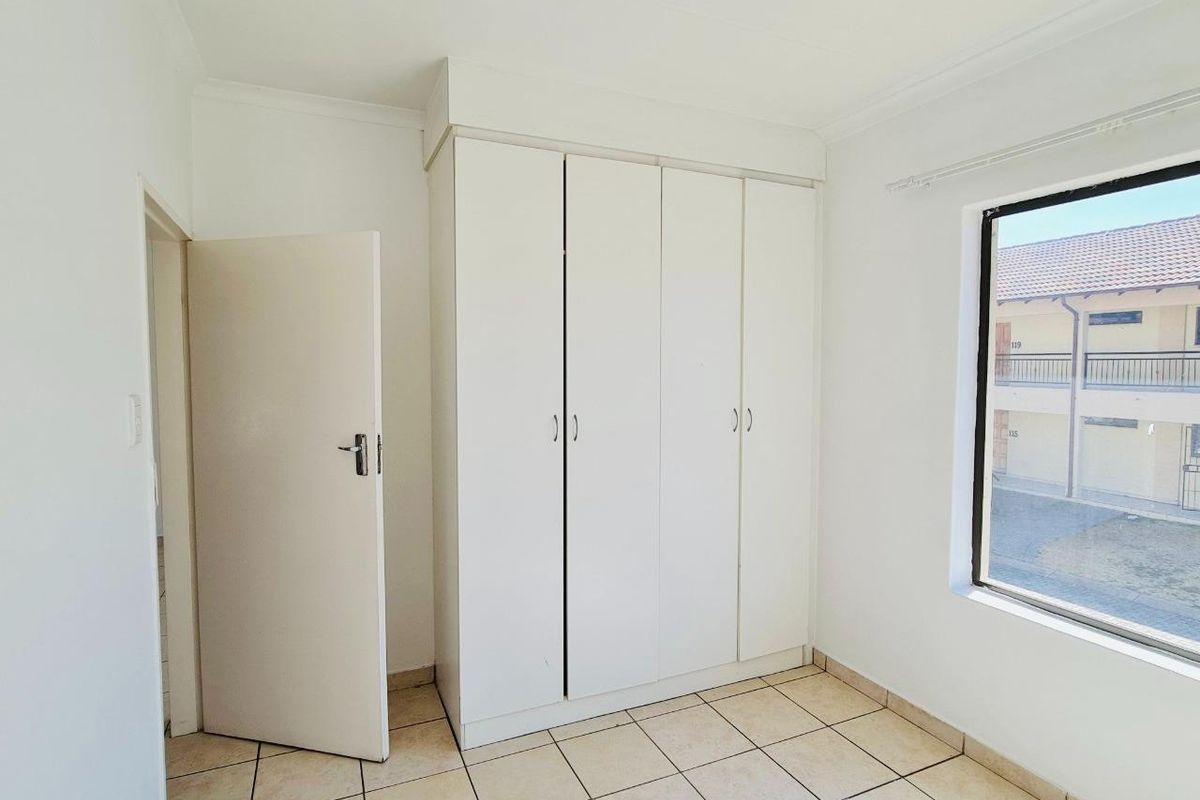 A bright, empty bedroom featuring built-in white cupboards, tiled flooring, and a window overlooking the apartment complex exterior.