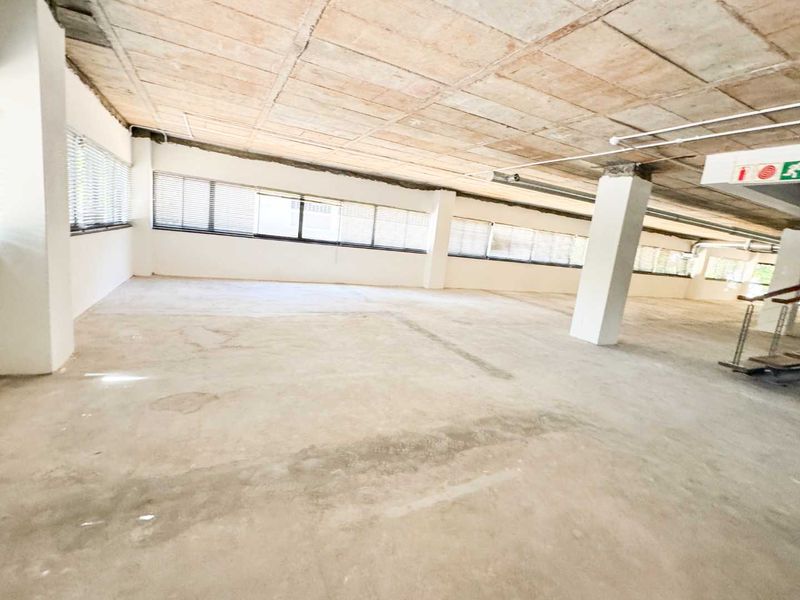 A large, open-plan office space featuring an exposed concrete ceiling, structural columns, and extensive window frontage with fitted blinds.