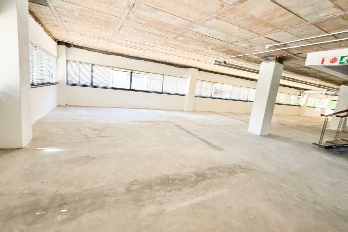 A large, open-plan office space featuring an exposed concrete ceiling, structural columns, and extensive window frontage with fitted blinds.