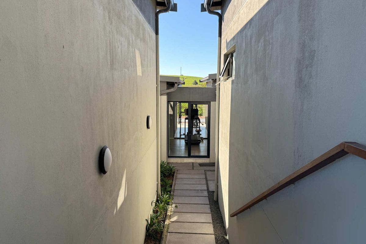 A narrow exterior walkway between building sections featuring paved path, wall-mounted lighting, and a wooden handrail leading toward a glass-enclosed entrance.