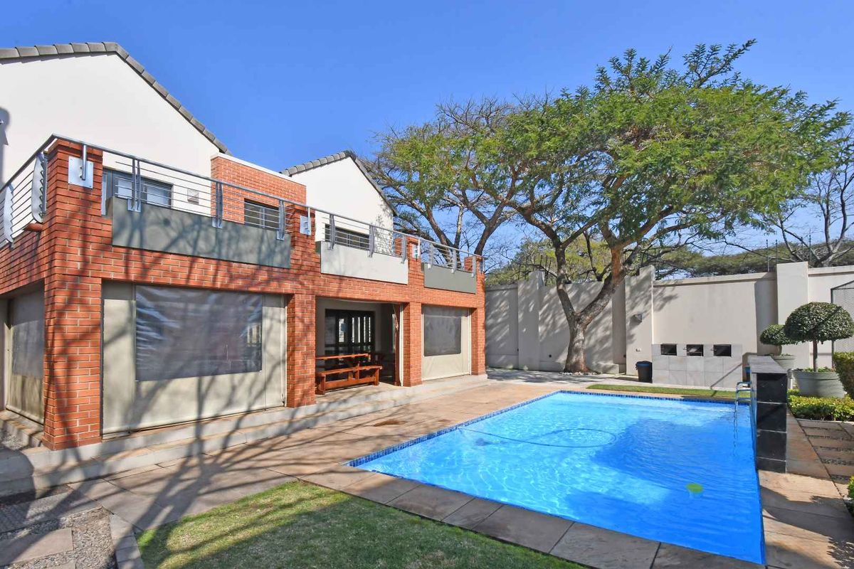 Rear exterior view featuring a swimming pool, paved patio area, built-in braai, and a brick facade with an upper-level balcony.