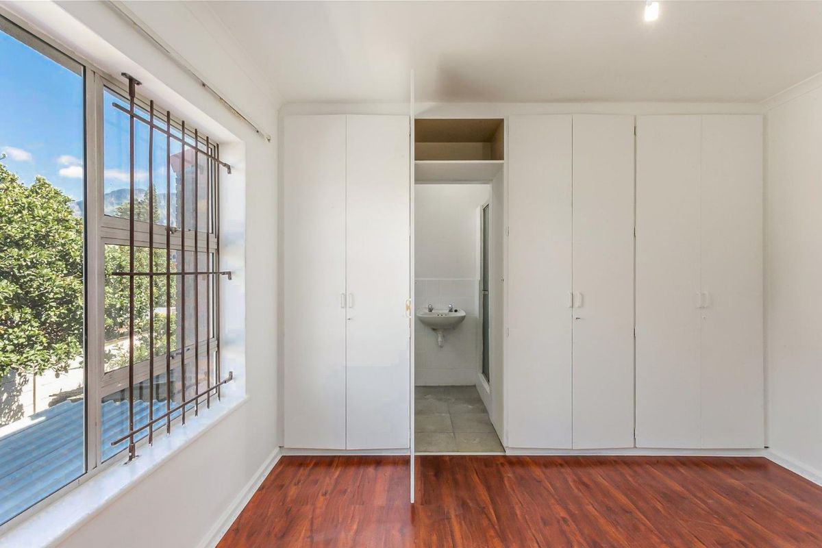 A bedroom featuring built-in cupboards, laminate flooring, and a window with security bars, leading to an en-suite bathroom area.
