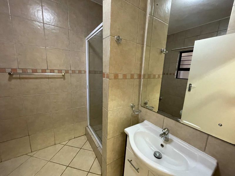 A functional bathroom featuring a tiled shower enclosure, a vanity basin with a wall-mounted mirror, and neutral wall and floor tiling.