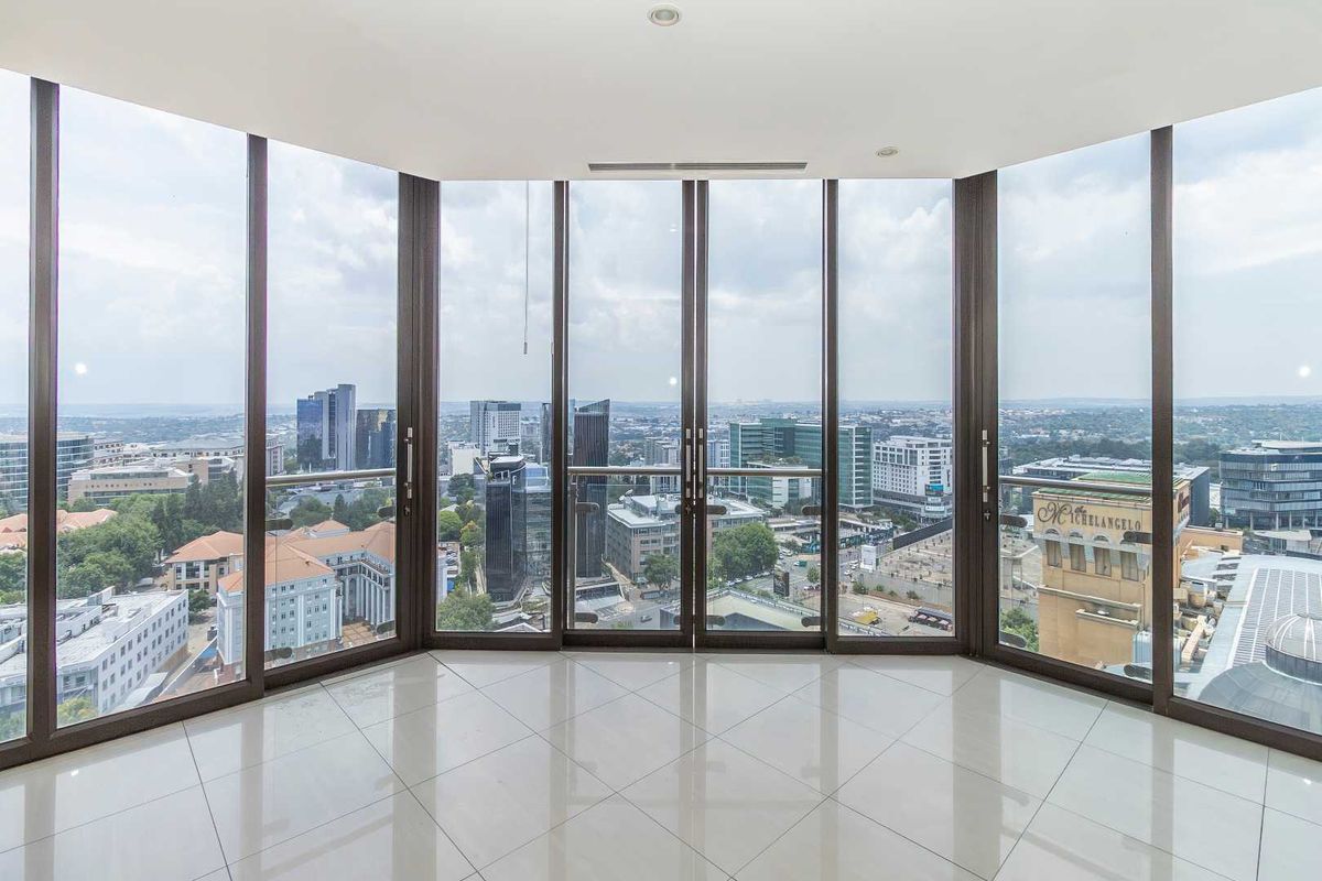 A bright, empty living area featuring floor-to-ceiling glass windows and doors that offer expansive panoramic city views and access to a balcony.