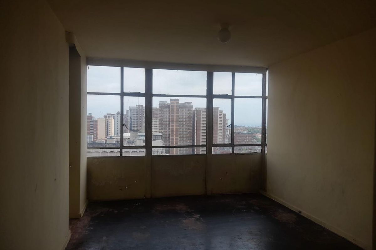 An empty room featuring large metal-framed windows with city views and worn flooring, offering a blank canvas for renovation.