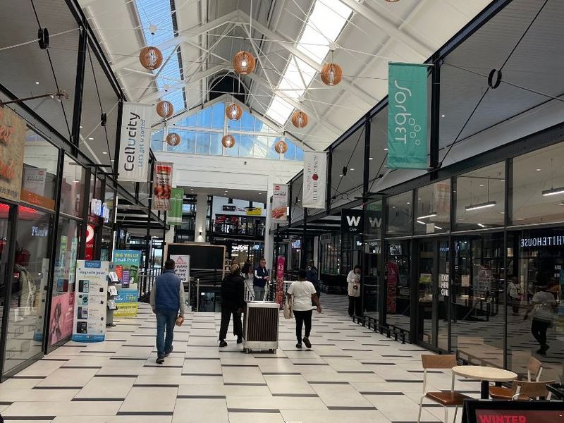 A bright, modern retail shopping centre corridor featuring high vaulted ceilings with skylights, tiled flooring, and glass-fronted retail units.