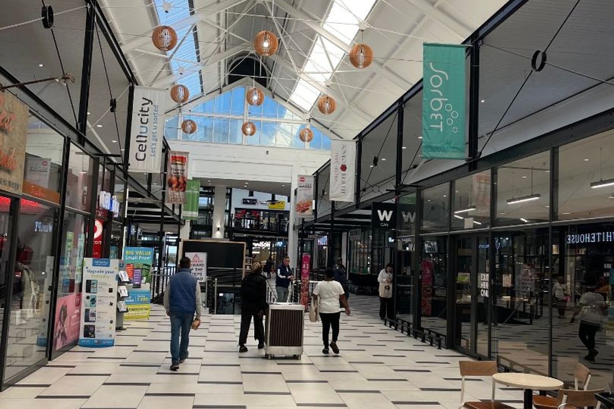 A bright, modern retail shopping centre corridor featuring high vaulted ceilings with skylights, tiled flooring, and glass-fronted retail units.